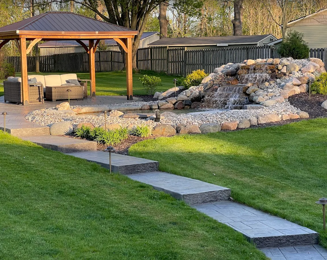Stamped concrete walkway leading to a paver patio, gazebo, and rock waterfall