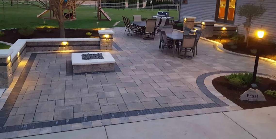 Large paver patio at dusk with built-in fire pit, seating wall, and landscape lighting